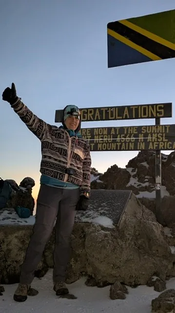A trekker beside the summit sign on Socialist Peak, Mount Meru, at 4,566m with the Tanzanian flag above